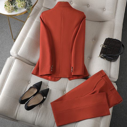 Ein gold-orangefarbener Blazer und eine dazu passende Hose von hinten betrachtet, präsentiert auf einer weichen Oberfläche mit schwarzen spitzen Absätzen und einer kleinen Handtasche daneben.